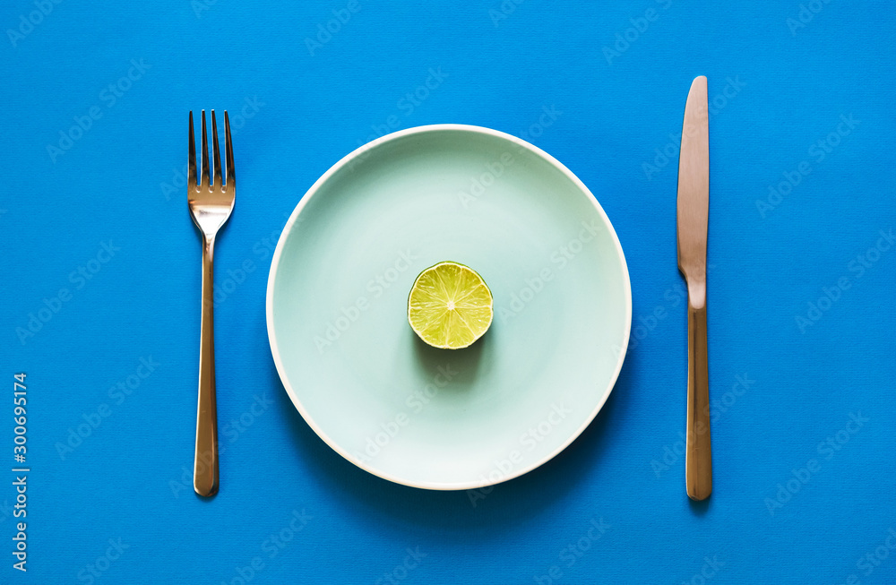A slice of fresh lime lying on color plate with fork and knife on blue background. Concept of vegan diet and citrus food. A slice of fresh lime lying on color plate with fork and knife on blue background. Concept of vegan diet and citrus food.