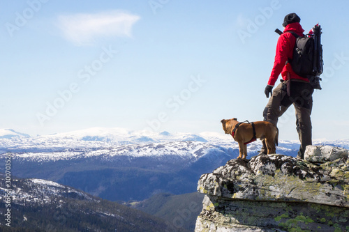 Man and dog standing on steep hillside and looking towards the snowy peaks of Jotunheimen in Norway. Staffordshire bullterrier, hiking, trekking, lifestyle, rondane and Norway concept.