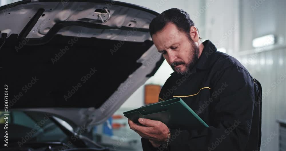 Smiling and happy mechanic man in a uniform while reading the documents from the map looking straight to the camera and feeling in a great mood in a auto service center Smiling and happy mechanic man in a uniform while reading the documents from the map looking straight to the camera and feeling in a great mood in a auto service center