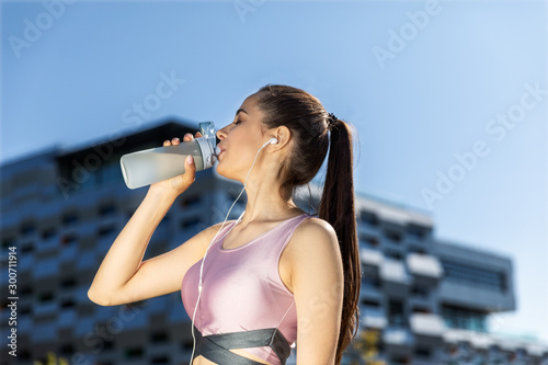 Girl with a ponytail is drinking from the sportive bottle in the earphones near the modern building