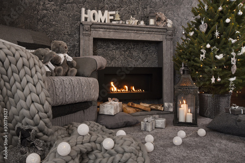 Modern cozy interior in gray shades with an armchair in front of fireplace and decorated Christmas tree. On the chair there are nice knitted blanket, cushions and toys. Candles, gifts and other Christ