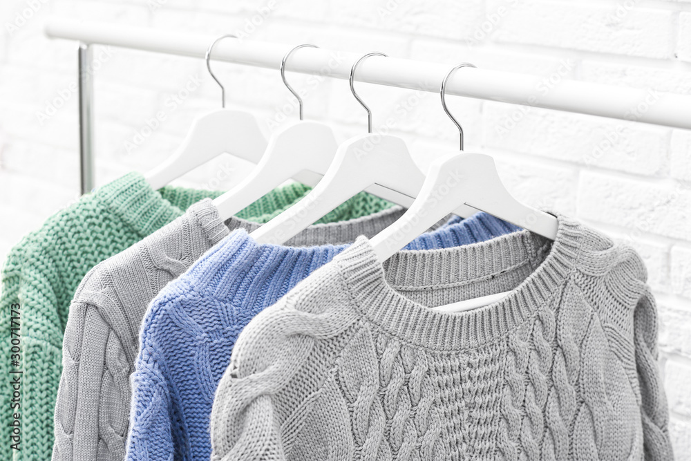 Collection of warm sweaters hanging on rack near brick wall, closeup - obrazy, fototapety, plakaty 