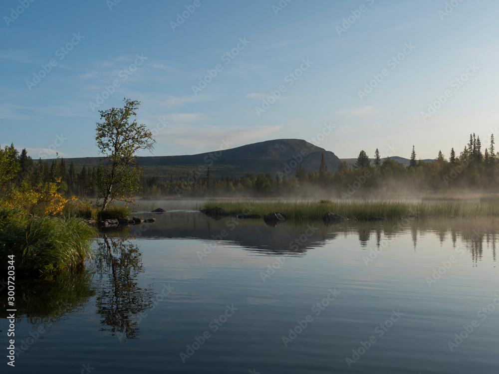 Beautiful morning over lake Sjabatjakjaure with haze mist in Sweden Lapland nature. Mountains, birch trees, spruce forest, rock boulders and grass. Sky, clouds and clear water.
