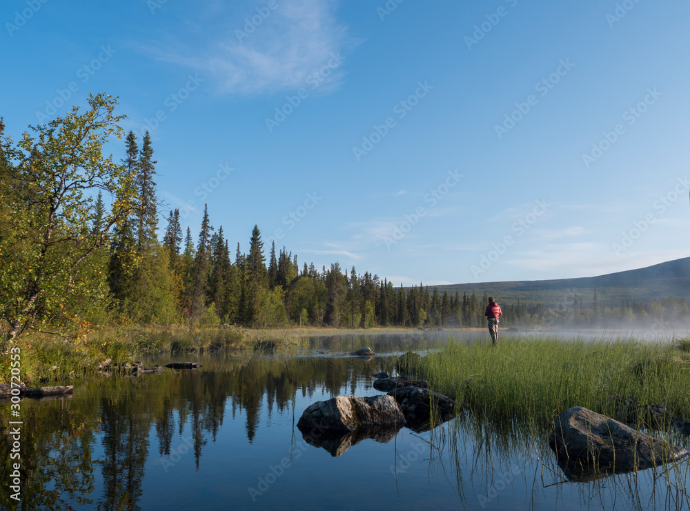 Fisherman man catching fish at lake Sjabatjakjaure in Beautiful sunny morning haze mist in Sweden Lapland nature. Mountains, birch trees, spruce forest. Blu sky