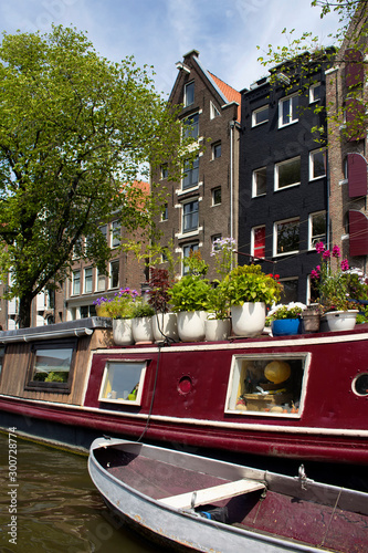 View of canal, a boat house with many flowers, trees and historical, traditional buildings showing Dutch architectural style in Amsterdam. It is a sunny summer day.