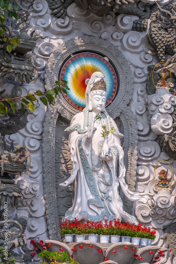 Da Nang, Vietnam - March 10, 2019: Chua An Long Chinese Buddhist Temple. Closeup of Gray stone Guan Yin statue. Red flowers at her feet, some green foliage. Chakra wheel behind her head.