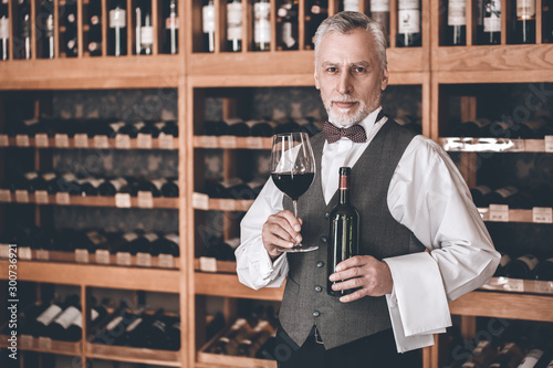 Sommelier Concept. Senior man standing with bottle and glass of wine smiling confident
