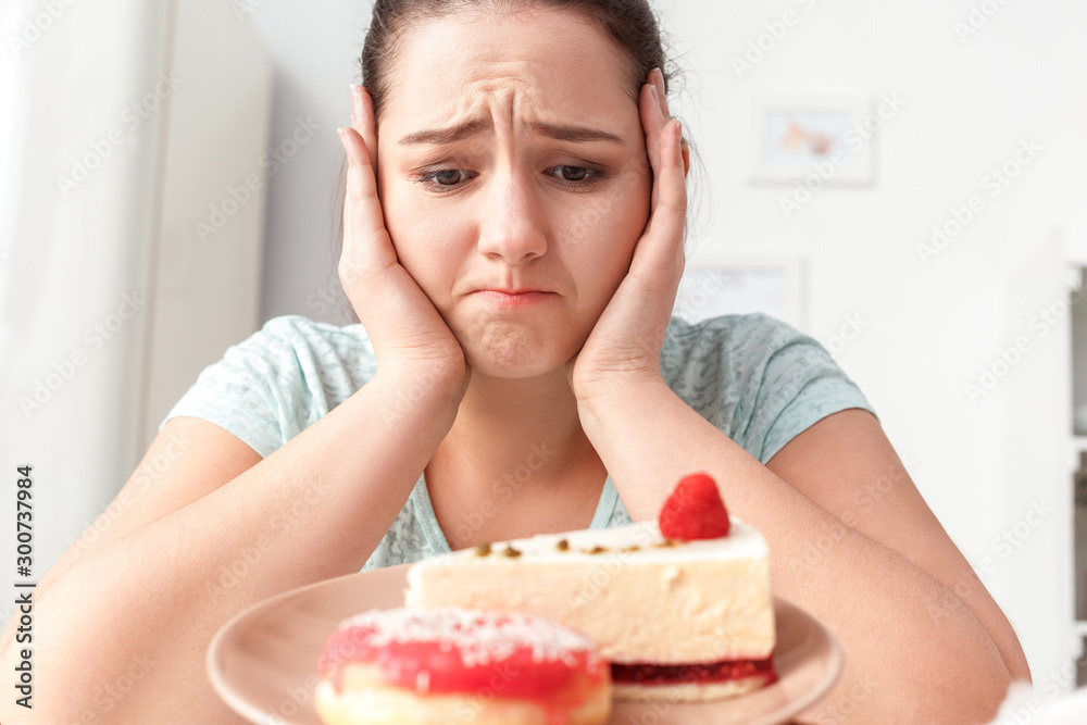 Junk Food. Chubby girl sitting at kitchen looking at plate with cake and doughnut unhappy close-up Junk Food. Chubby girl sitting at kitchen looking at plate with cake and doughnut unhappy close-up