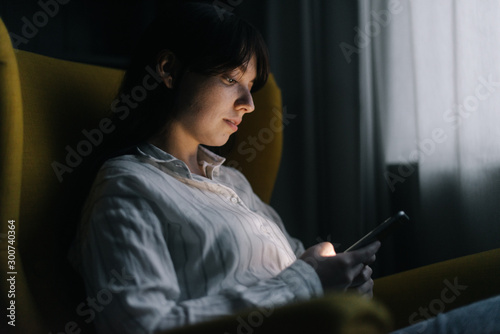 Beautiful young woman is using phone in dark room. Attractive girl sits on soft yellow chair. Modern evening pastime. © dikushin