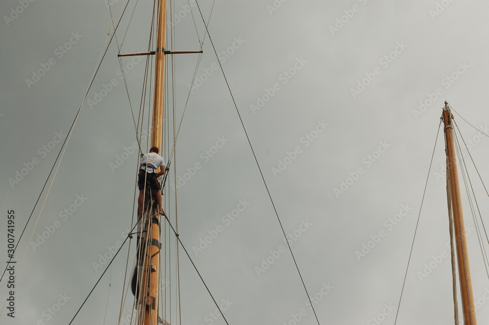 Mast of wooden sailing boat, spreaders, shrouds. A sailor is climbing up on a mast. A cloudy sky. Race at Saint-Tropez, Cote d'Azur, France. Mast of wooden sailing boat, spreaders, shrouds. A sailor is climbing up on a mast. A cloudy sky. Race at Saint-Tropez, Cote d'Azur, France.