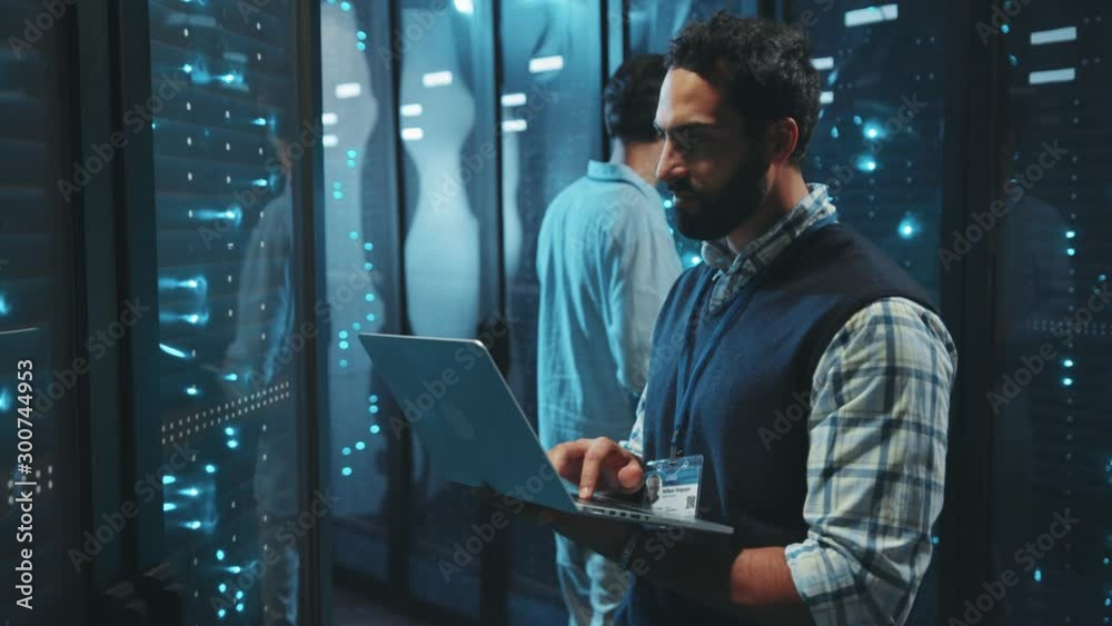 Bearded server specialist using computer laptop running diagnostics on server rack cabinets inspecting digital room of data center.