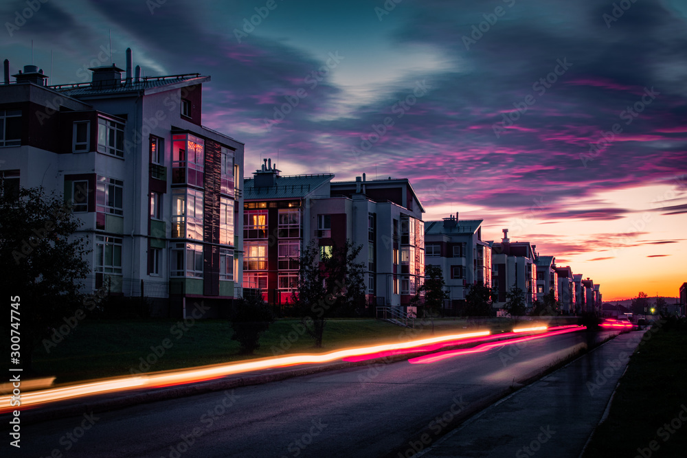Obraz premium Light trails of passing cars in front of houses during sunset with purple clouds