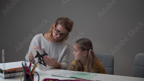 Intelligent Caucasian woman explaining Rorschach test card to the little girl. Female Caucasian psychologist diagnosing child at her office. Pretty kid passing psychological test at school. photo