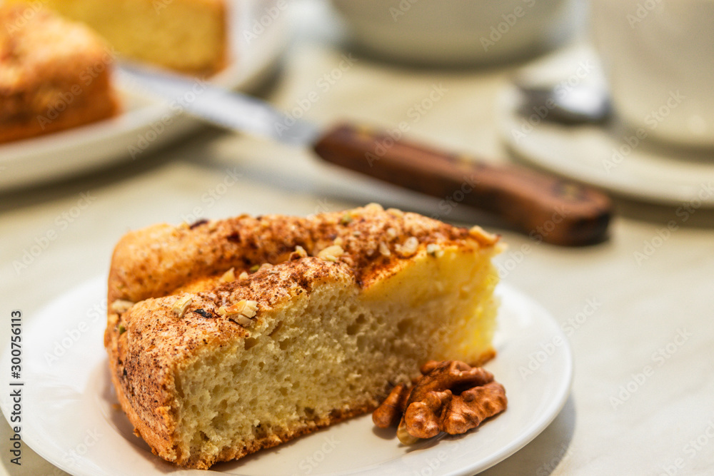 A piece of cake with apples, chopped walnuts and cinnamon lies on a white plate next to a brown knife, close-up