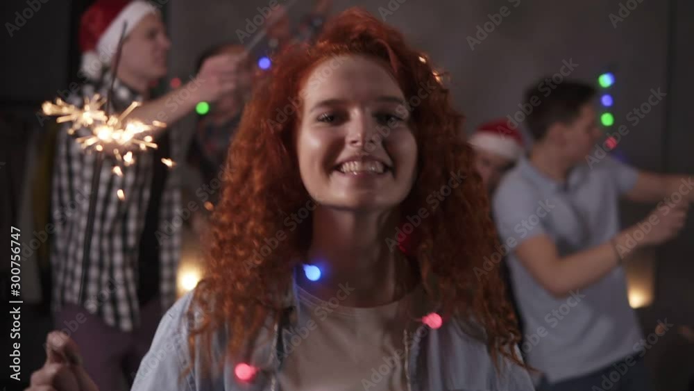 Precious curly redhaired girl posing for camera - dancing and smiling, waving her bengal light while her friends dancing on the blurred background in decorated room. Friends celebrating new year Precious curly redhaired girl posing for camera - dancing and smiling, waving her bengal light while her friends dancing on the blurred background in decorated room. Friends celebrating new year