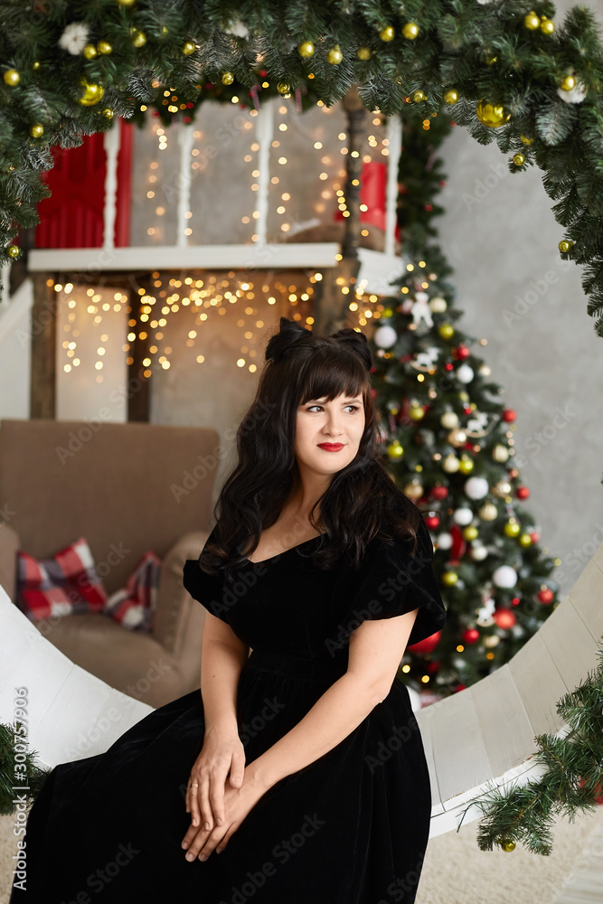 Young plus size model woman with bright makeup and modish hairstyle in a black dress posing in interior decorated for New year. Fashionable plus size girl in trendy outfit posing near Christmas tree Young plus size model woman with bright makeup and modish hairstyle in a black dress posing in interior decorated for New year. Fashionable plus size girl in trendy outfit posing near Christmas tree