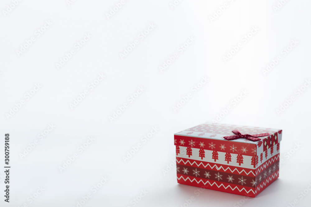 Obraz premium A red Christmas box with gifts and a bow stands on a white background with place for postcard text on the right. On the box are Christmas trees and snowflakes.