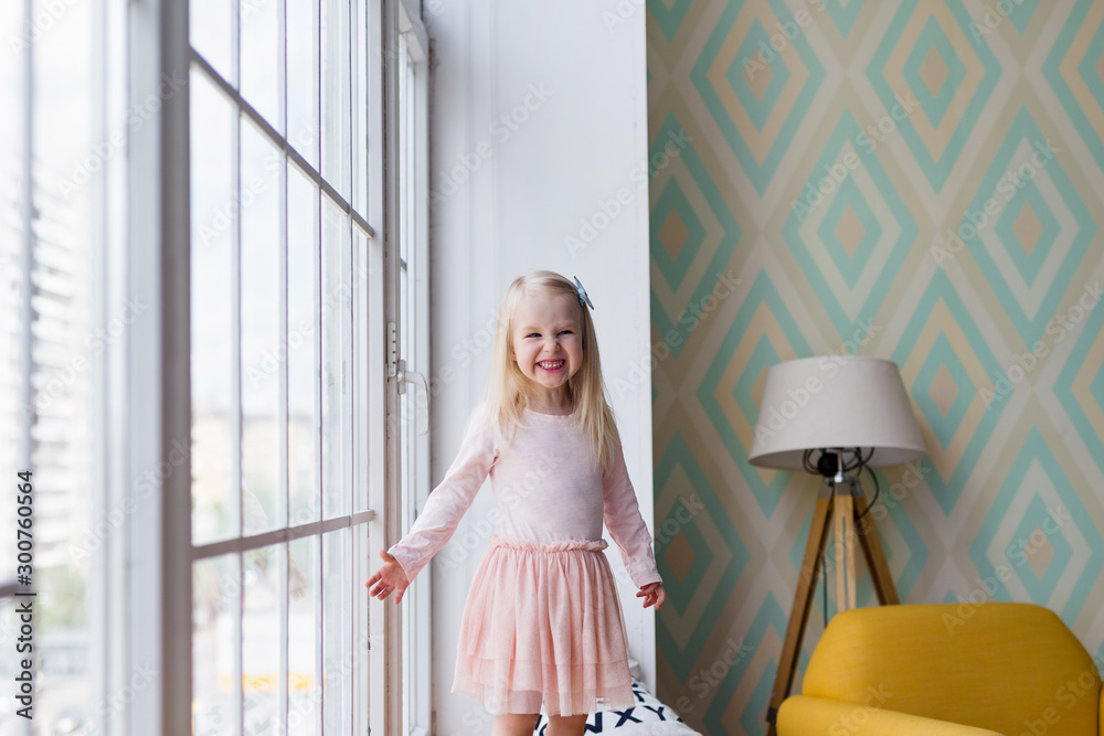Happy little girl smiling while standing in front of window in cozy room. Funny 3 years old girl laughing in front of window