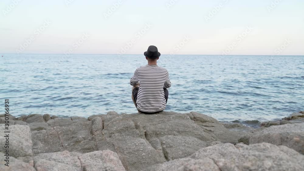 Caucasian man in hat, shorts and striped shirt sitting by the seashore, looking at sunset. Meditation and observation of ocean view. Sea cost, vacation and calm , relaxed evening at the ocean. Caucasian man in hat, shorts and striped shirt sitting by the seashore, looking at sunset. Meditation and observation of ocean view. Sea cost, vacation and calm , relaxed evening at the ocean.