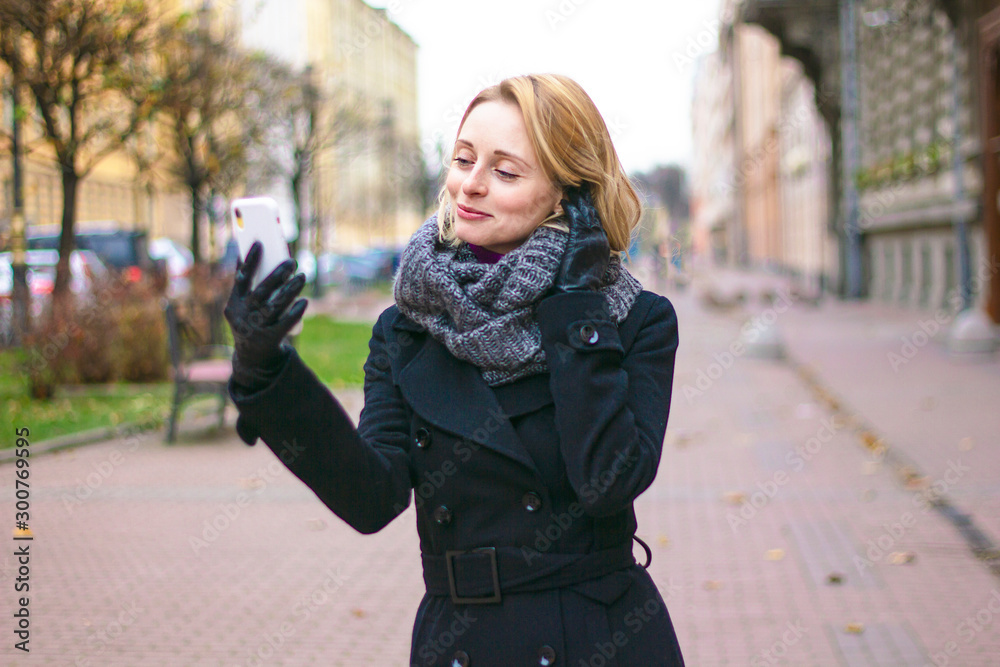 Fototapeta premium Satisfied woman takes a selfie. Happy girl talking on a cell phone with a face camera. Lady is phoning in the park on a cloudy day outside