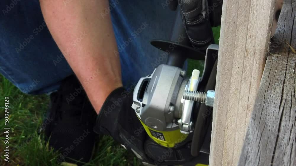 A man is using a grinder a metal screw and bolt that is attached to a wooden fence. Sparks are seen flying off the screw. A man is using a grinder a metal screw and bolt that is attached to a wooden fence. Sparks are seen flying off the screw.
