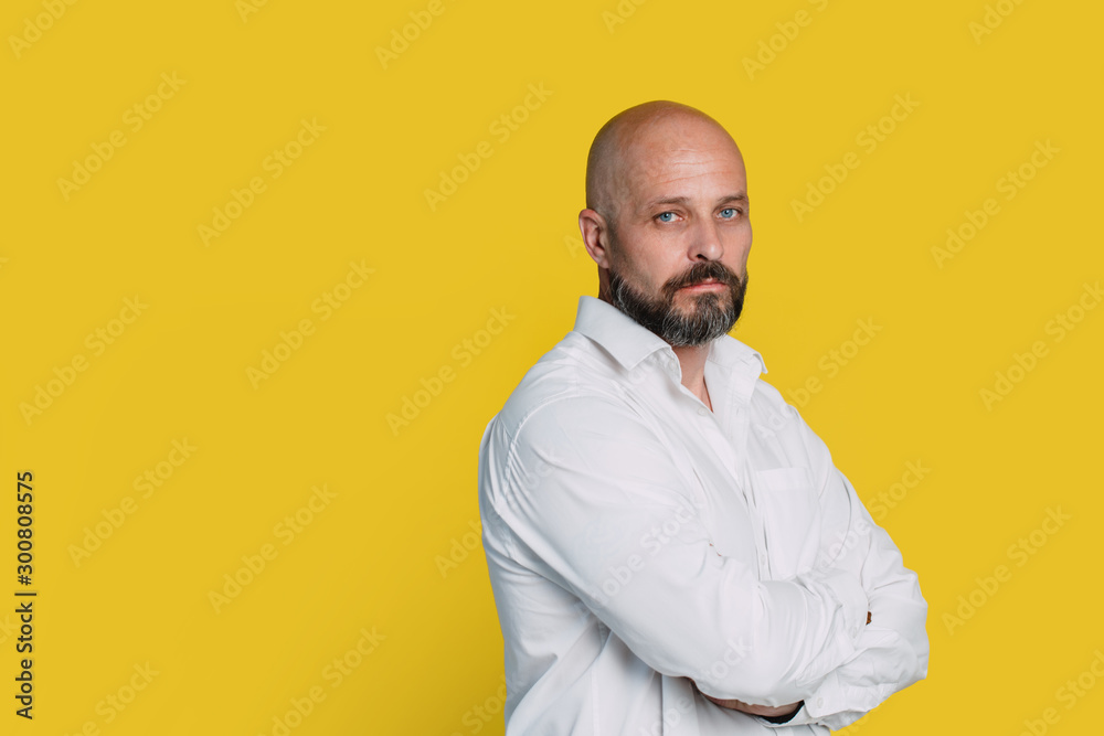 Serious middle-aged man with folded arms and a deadpan expression posing in front of a yellow background with copy space