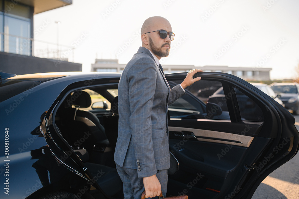 Portrait of a handsome businessman in sunglasses standing near the car outdoors in front of the modern building facade
