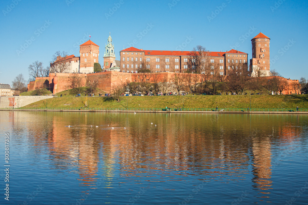 The view of Wawel castle in Krakow city with reflection in the water, ducks floating on the foreground, group of tourist walking around castle in sunny winter day