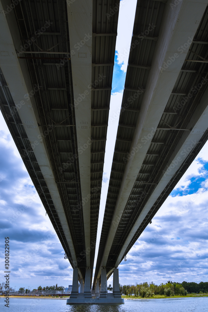 Large bridge over the river, view from below. On the background of contrasting clouds and blue sky. Large bridge over the river, view from below. On the background of contrasting clouds and blue sky.