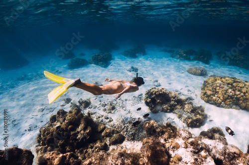 Freediver girl with yellow fins glides over sandy bottom with fishes in ocean