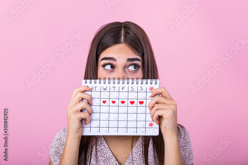 Portrait of a funny young girl in hiding behind a menstrual periods calendar and looking away at copy space isolated over pink background. Female Period calendar photo