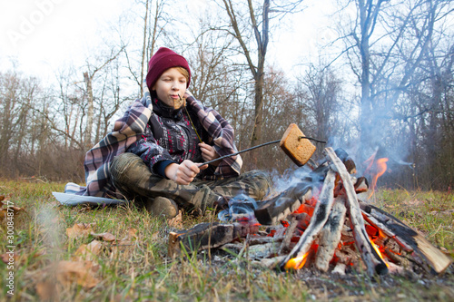 A boy sits by a fire in a forest glade and grills a barbecue.