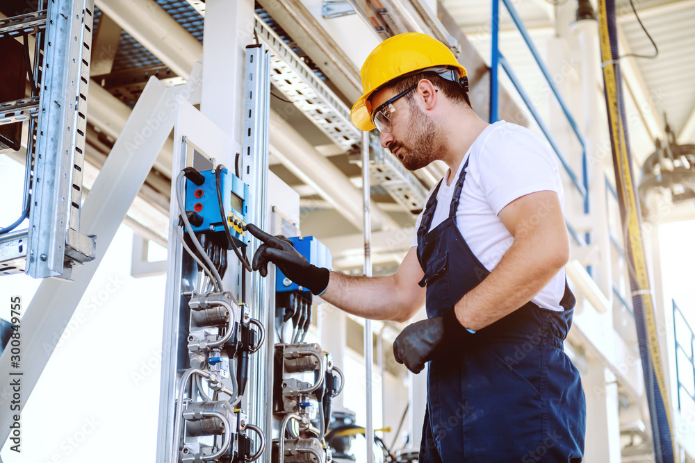 Side view of handsome caucasian worker in overall and with helmet on head pressing button on dashboard. Oil production. Side view of handsome caucasian worker in overall and with helmet on head pressing button on dashboard. Oil production.