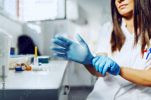 Cropped picture of caucasian attractive female laboratory assistant in white uniform sitting in lab and putting sterile rubber gloves.