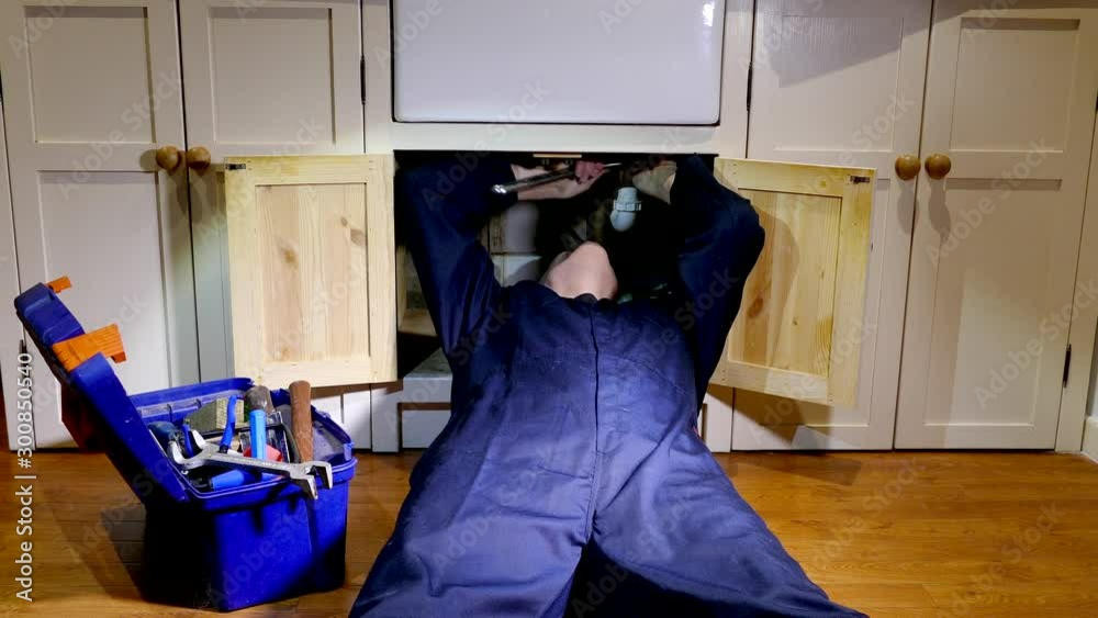POV shot at ground level of a man plumber wearing blue overalls, lying on his back on the floor and using a large wrench to tighten a nut in the pine cupboard beneath a kitchen sink.