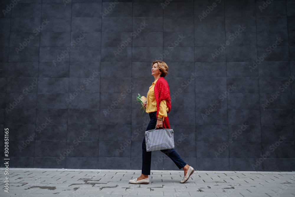 Side view of charming caucasian fashionable senior woman carrying bag and groceries while passing by gray wall.