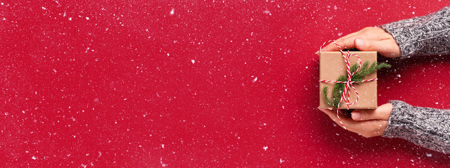 Female's hands in pullover holding Christmas gift box decorated with evergreen branch on red background with snow. Christmas and New Year banner.