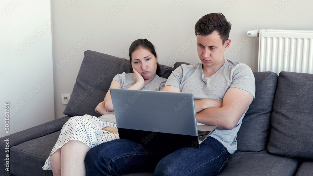 Couple is watching bored film and falling asleep. Young man and woman watch video on laptop sitting on sofa. They are in their modern flat. Couple is watching bored film and falling asleep. Young man and woman watch video on laptop sitting on sofa. They are in their modern flat.