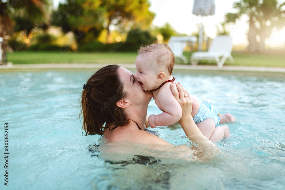Attractive dedicated Caucasian brunette kissing her loving 6 months old son while standing in swimming pool. First time at pool concept.