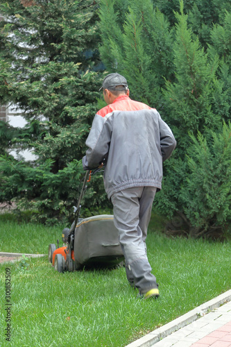 An adult man in uniform with an automatic lawn mower cuts a lawn in the city park on a summer day