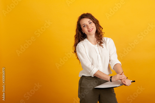 Beautiful young girl with a clipboard and a pencil smiles and looks at the camera standing isolated on yellow background.
