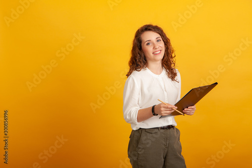 Beautiful young girl with a clipboard and a pencil smiles and looks at the camera standing isolated on yellow background.
