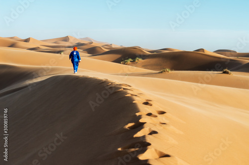 Traditional dressed Moroccan man with turban walks on a sand dune in the Sahara desert.