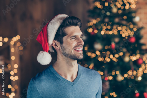 Photo of funky cute cheerful handsome man in santa headwear smiling toothily with bristle lights ornament garland illumination behind him with fir tree