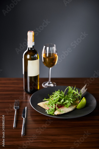 tasty restaurant fish steak with lime and arugula on wooden table near cutlery and white wine on black background