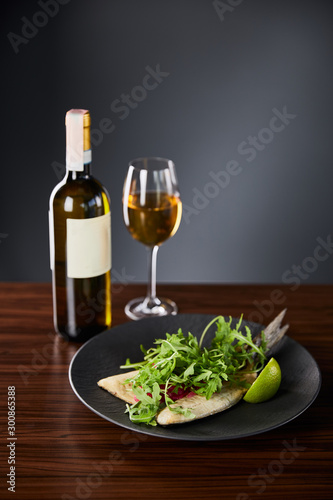 tasty restaurant fish steak with lime and arugula on wooden table near white wine on black background