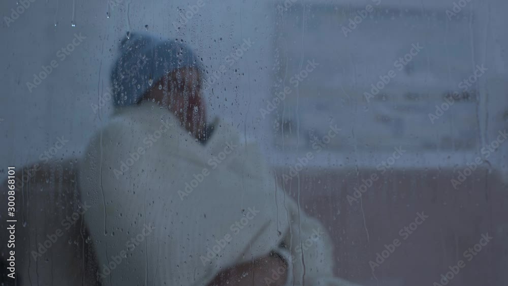 sick man sitting near window glass with raindrops and sneezing into napkin