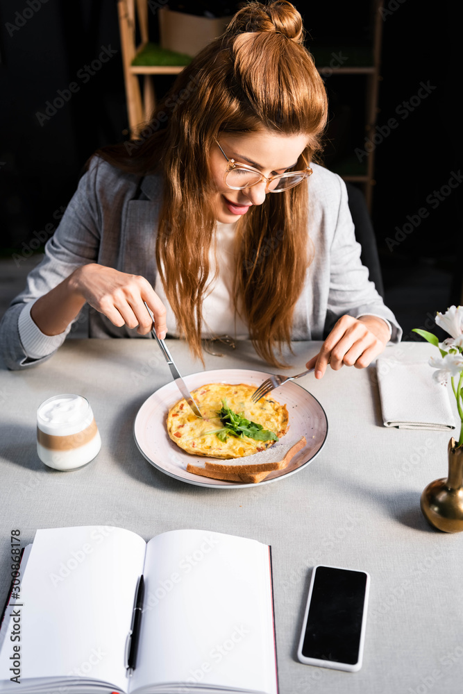 redhead girl eating omelet for breakfast with coffee in cafe with empty notepad and smartphone