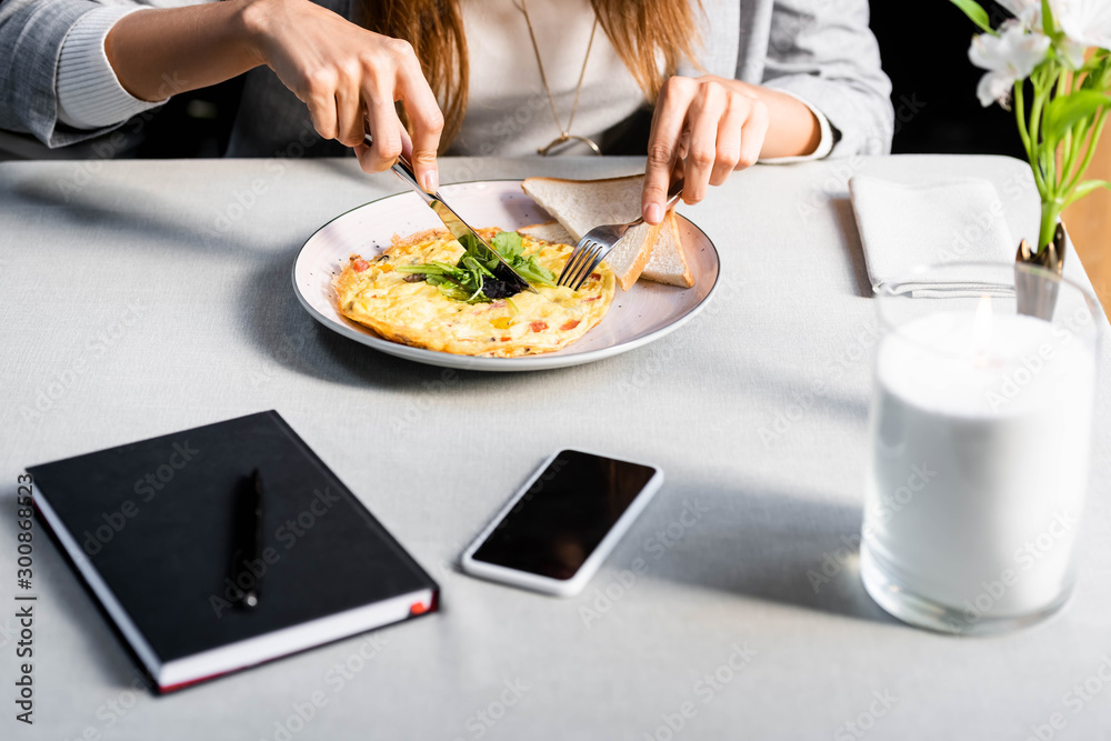 cropped view of woman eating omelet for breakfast in cafe with notepad and smartphone