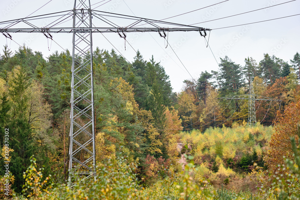 Φωτογραφία High-voltage powerlines and high-voltage pylons transporting  electric power in a lane through a beautiful autumnal forest
